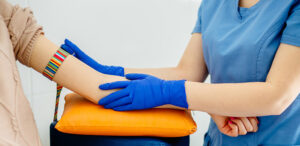 A clinician in scrubs wearing gloves preparing a patient's arm for a blood test, with a tourniquet applied above the wrist