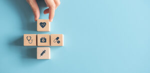 A hand arranging wooden blocks printed with medical icons — a heart, stethoscope, first-aid kit, pills and syringe — into a cross shape